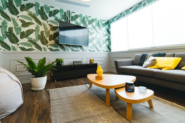A modern living room with a tropical leaf wallpaper, a gray sofa with yellow pillows, a TV, a plant, and a wooden coffee table on a rug.