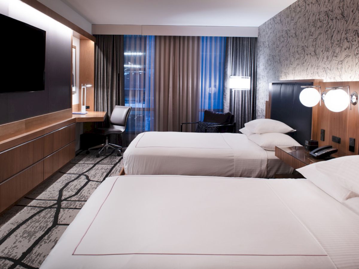 A modern hotel room with two beds, a wall-mounted TV, a desk with a chair, and a lamp beside the window with curtains drawn.