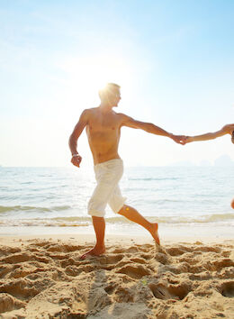 A man and woman holding hands are running on a sandy beach near the ocean under a bright sky.