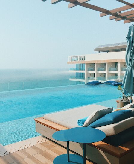 A luxurious poolside setup with lounge chairs, blue umbrellas, and an infinity pool overlooking a calm sea under clear skies.