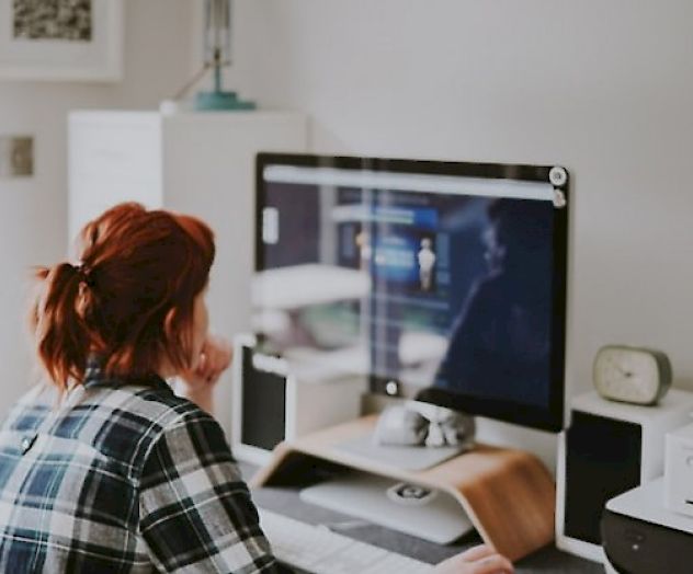 A person with red hair is sitting at a desk, working on a computer in a home office setting, with various items around.