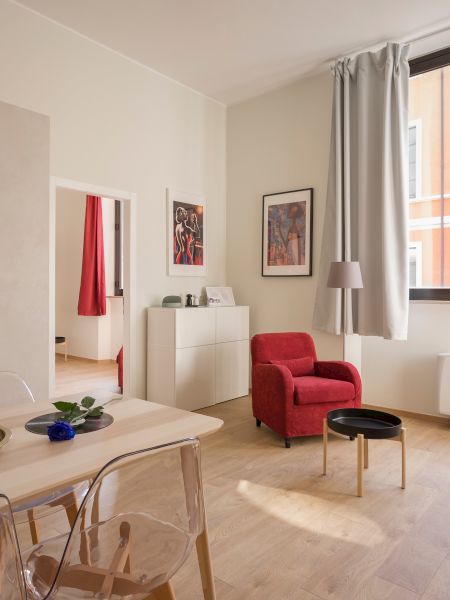 A modern living space with a kitchen, dining table, red armchairs, artwork on the walls, and a window with a view of neighboring buildings.