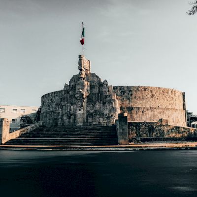 A large stone monument with intricate carvings and steps leading up to it, topped with a flag, set against a backdrop of trees and a building.