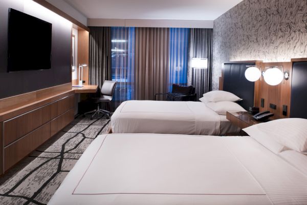 A modern hotel room with two beds, a flat-screen TV, a desk with a chair, and artistic decor on the walls. The room is well-lit and stylish.