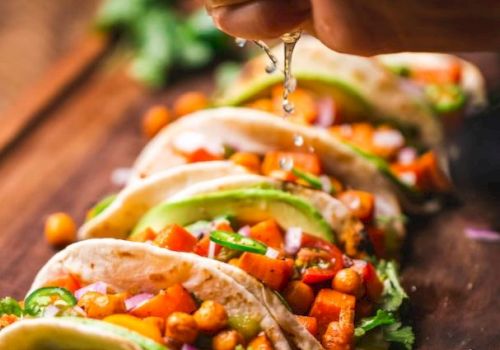 The image shows a hand squeezing a lime over a row of colorful tacos filled with vegetables and chickpeas on a wooden surface.