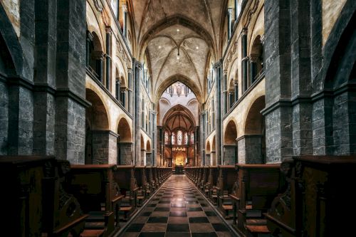 This image depicts the interior of a grand cathedral featuring high vaulted ceilings, arched columns, and rows of wooden pews lining a central aisle.