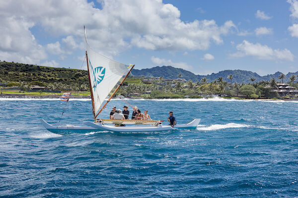 A traditional sailing boat with several people is gliding over bright blue waters against a backdrop of lush hills and palm trees.