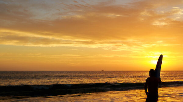 A person standing on the beach holding a surfboard is silhouetted against a vibrant sunset sky with clouds reflecting on the calm ocean water, ending the sentence.