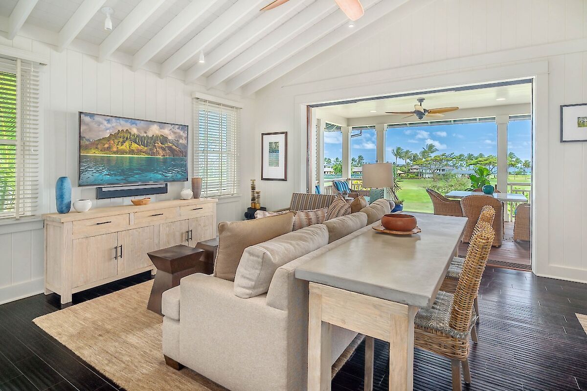 Cozy, airy living room with a beige sofa, rustic coffee table, and a TV on a white cabinet; sliding glass doors open to a sunny patio and ocean view.