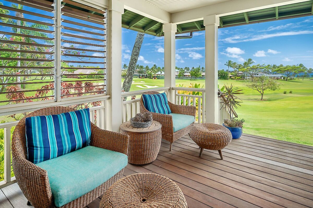 A sunlit screened porch with wicker chairs, blue cushions, and round coffee tables overlooking a green lawn and palm trees.