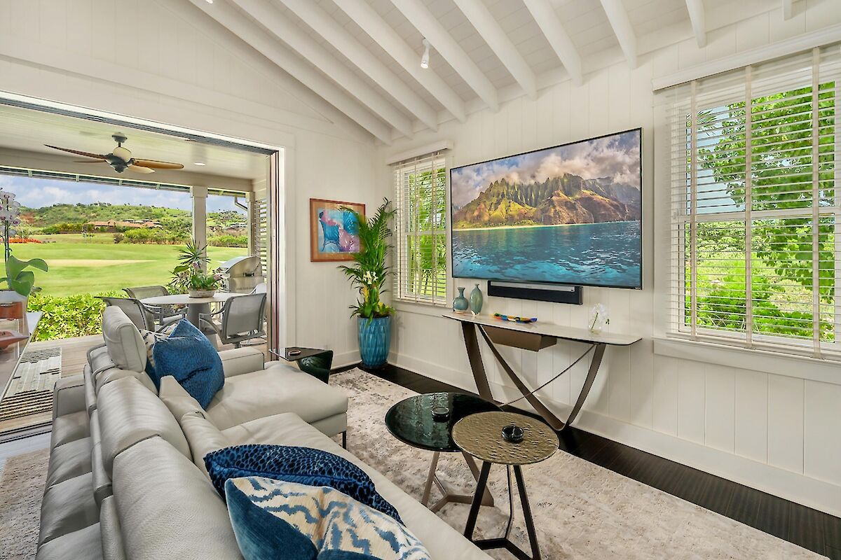 A bright coastal living room with white walls, a large TV above a console, gray sectional sofa, glass-top tables, and windows unveiling a green, sunny outdoor view.