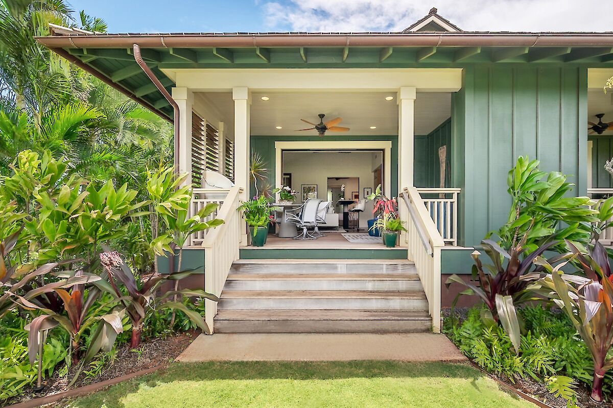 A tropical-style house with a covered porch, greenery surrounding, and a seating area visible from the front steps, inviting and sunny.