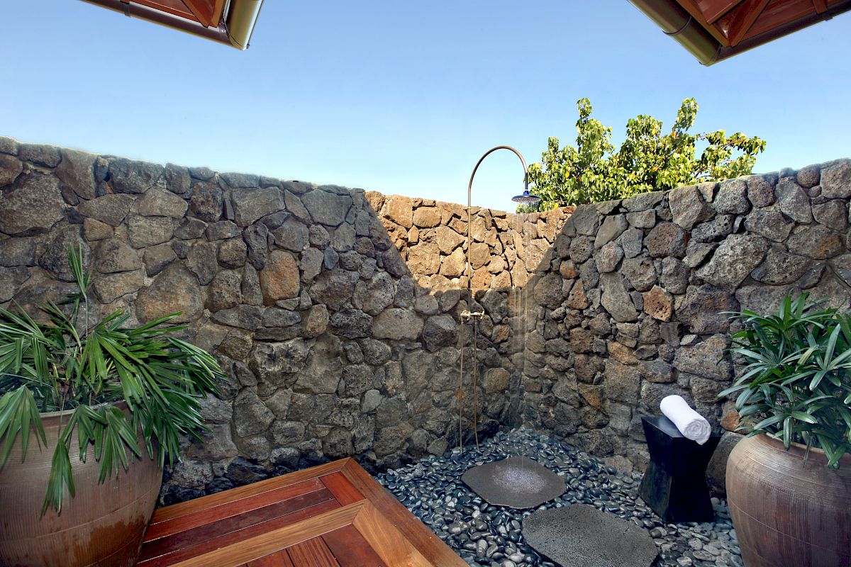 The image features a serene outdoor space with stone walls, tropical plants, and wooden decking under a clear blue sky.