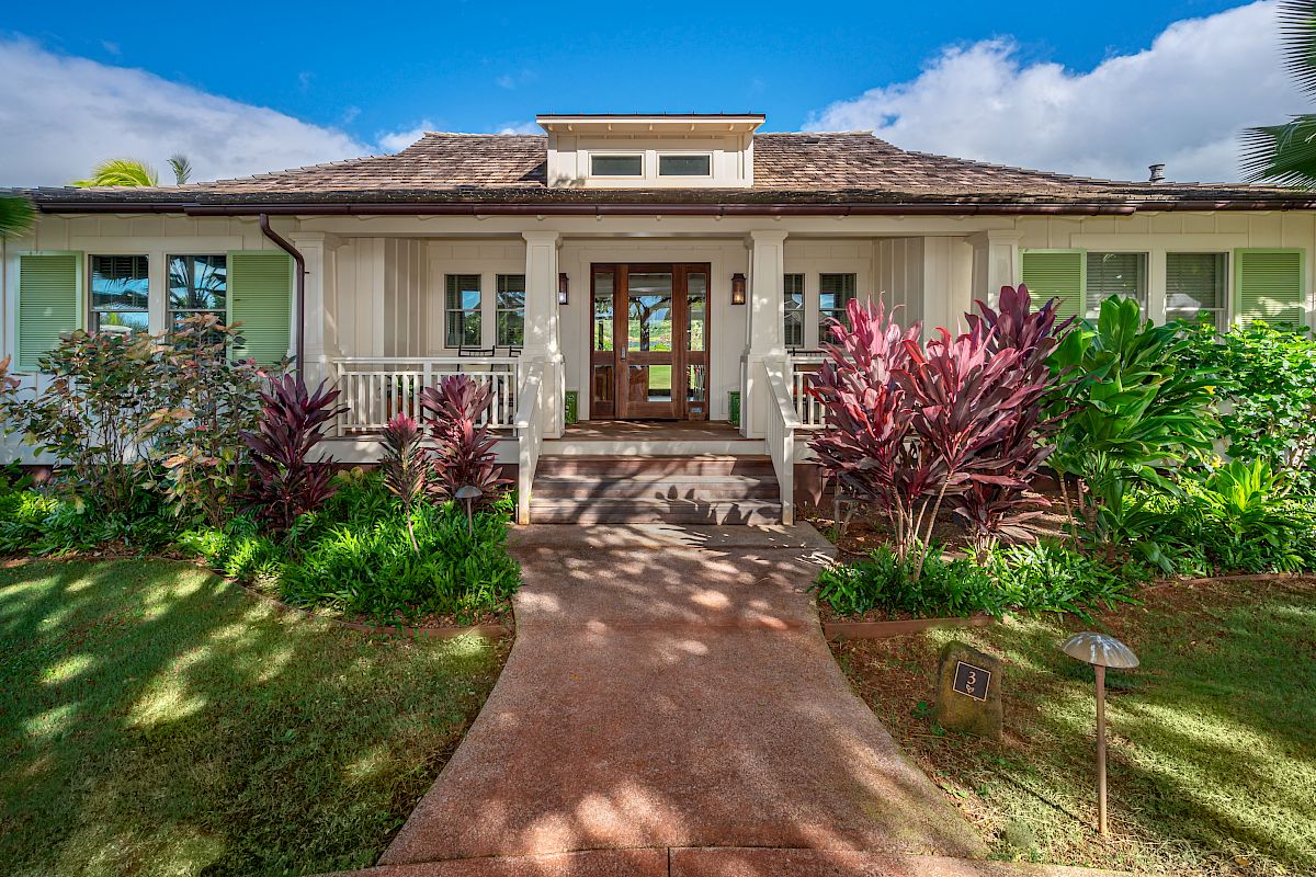 The image shows a charming house with a welcoming front porch, lush greenery, and colorful plants around the pathway.