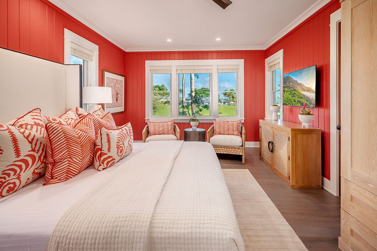 A bright bedroom with red walls, a bed with patterned pillows, a dresser, TV, chairs, and large windows looking out to greenery ends the sentence.