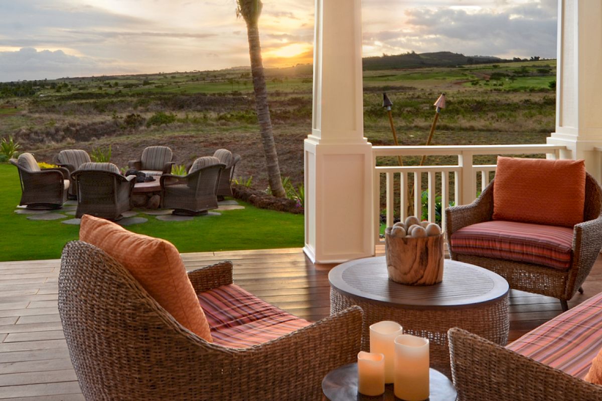 A cozy patio with wicker furniture and orange cushions overlooks a lush lawn with seating and a scenic landscape beyond, under a setting sun.