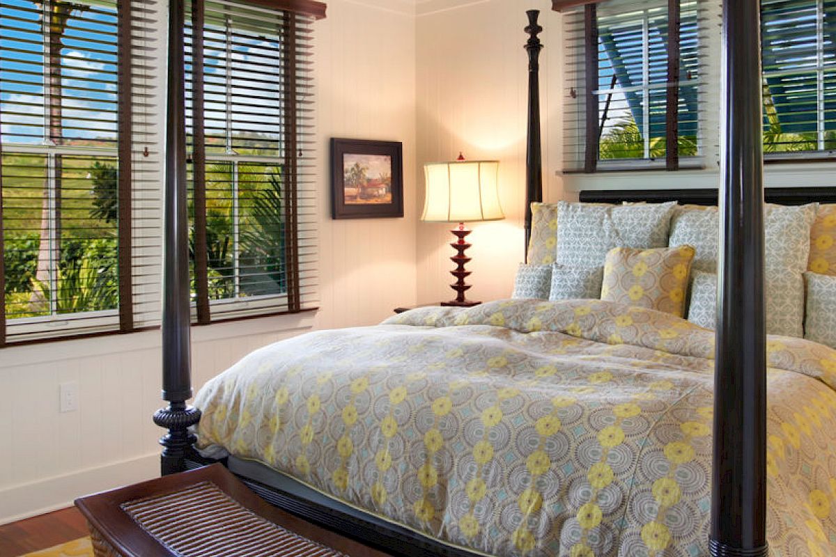A cozy bedroom with a four-poster bed, yellow and gray bedding, two nightstands with matching lamps, windows with blinds, and decorative artwork ending the sentence.