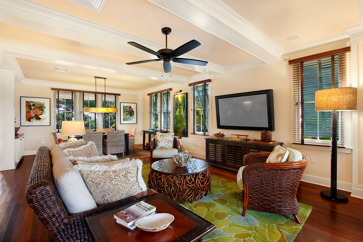 The image shows a cozy living room with comfortable seating, a TV, a ceiling fan, and bright decor, featuring natural light.