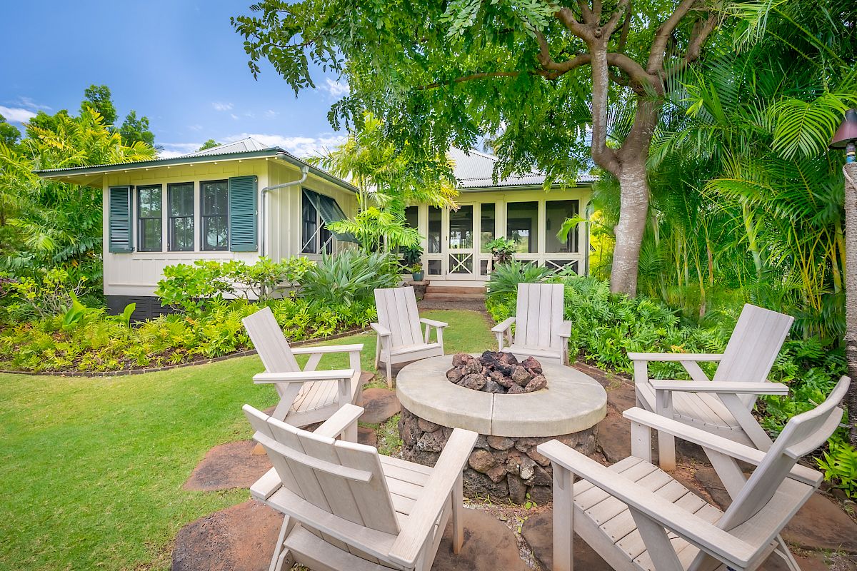 The image shows a cozy garden with a fire pit surrounded by wooden chairs, and a house in a lush, green setting.