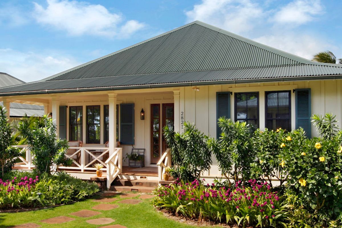 A charming white house with a green metal roof, surrounded by lush greenery, colorful flowers, and a well-maintained garden pathway.