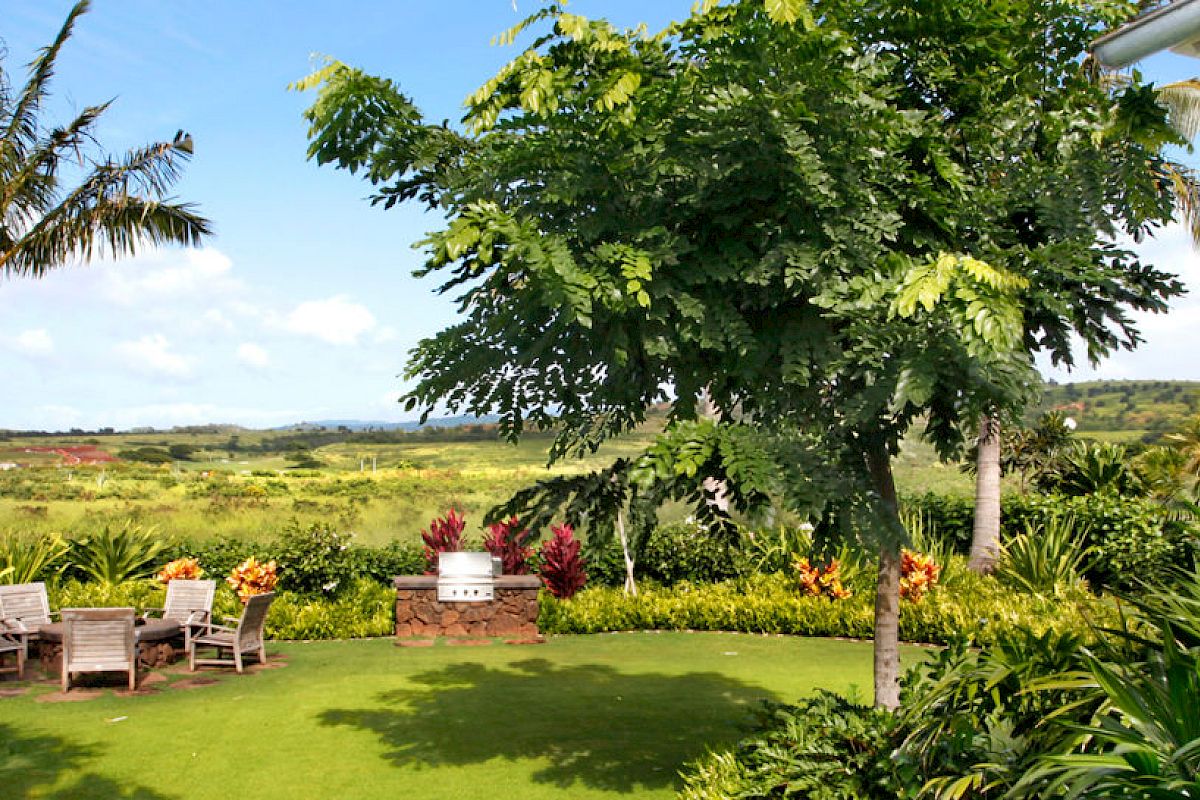 A lush garden with palm trees, various greenery, and outdoor seating, with a scenic landscape and a wide sky view in the background.