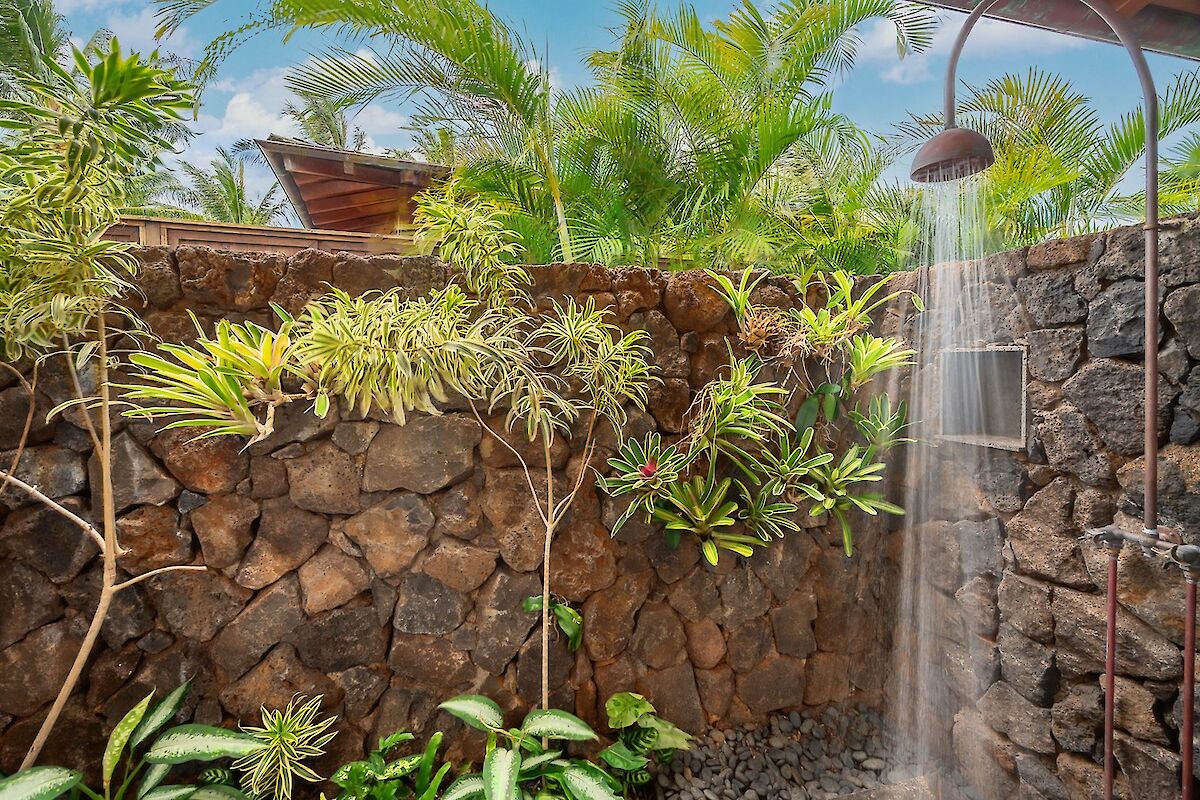A tropical outdoor shower setup with a stone wall, lush green plants, and a rainfall shower head creating a mist.
