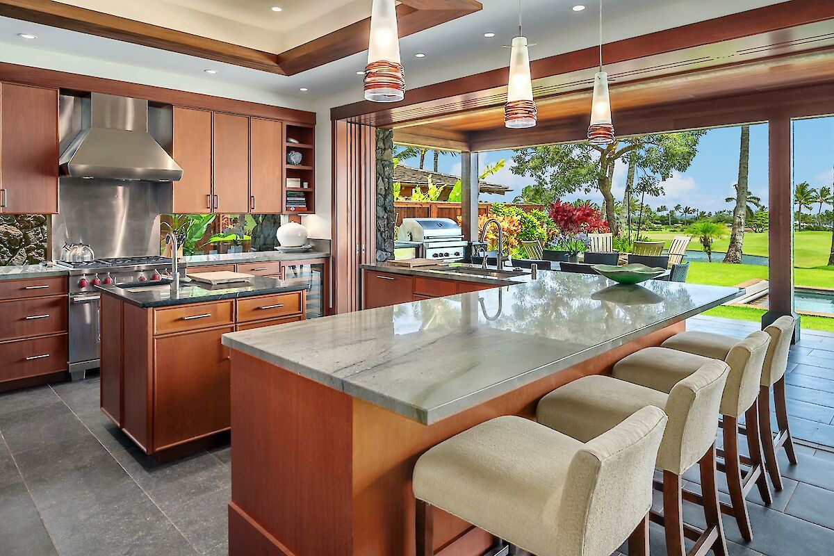 A modern kitchen with a large marble island, wooden cabinets, stainless steel appliances, and a bright open view to a lush backyard.
