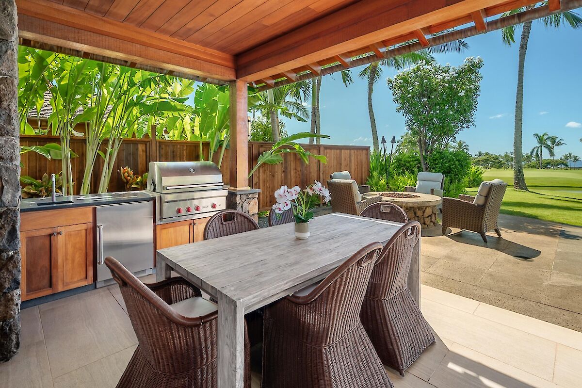 Outdoor kitchen and dining area with a wooden table, wicker chairs, built-in grill, lush tropical plants, and a sunny lawn backdrop.