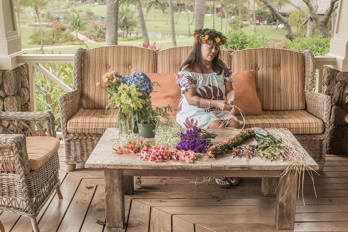 A woman sits on an outdoor sofa, making leis with various flowers and plants on a wooden table in a garden setting, wearing a floral crown and dress.