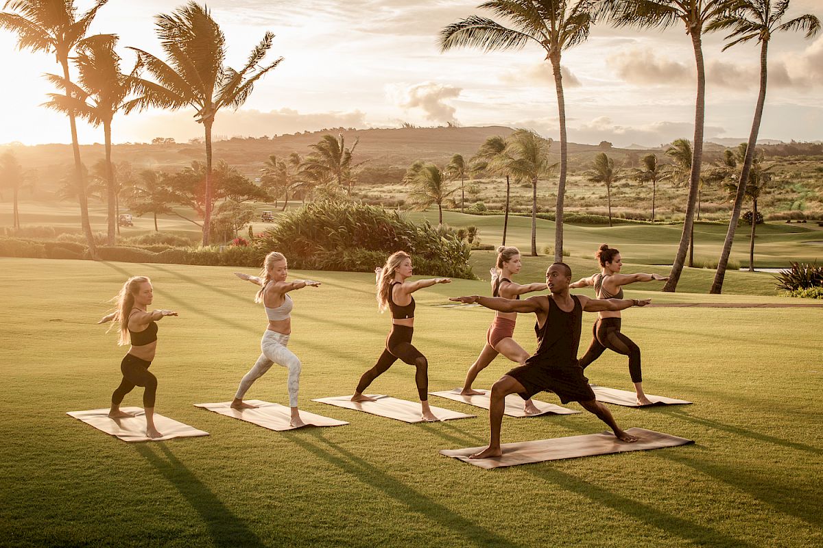 A group of people practicing yoga outdoors with mats on a grassy field during sunset, surrounded by palm trees.