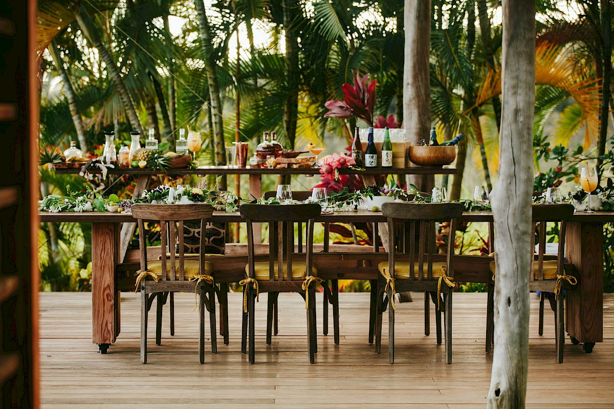 An outdoor dining setup with wooden chairs around a long table, surrounded by lush greenery and decorated with flowers and tableware, ending the sentence.