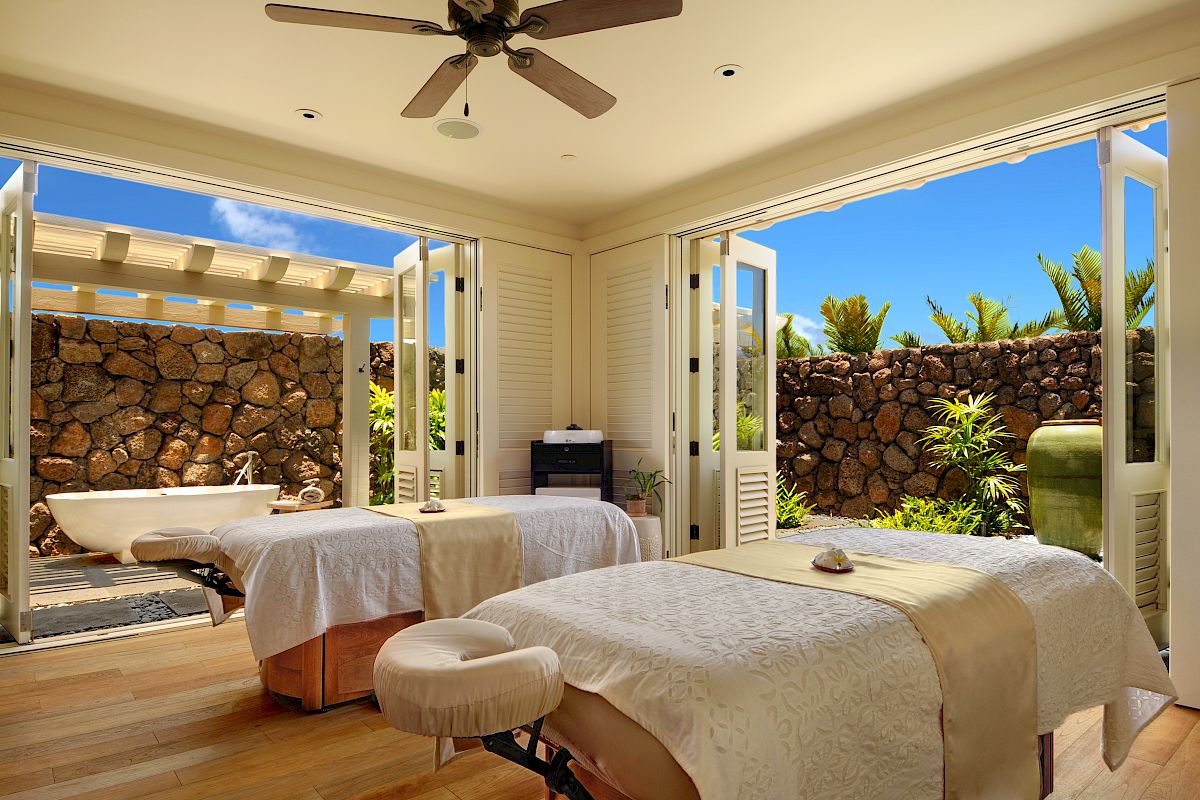 A luxurious spa room with two massage tables, open French doors, and an outdoor area with a bathtub and stone wall under a clear blue sky, ending the sentence.