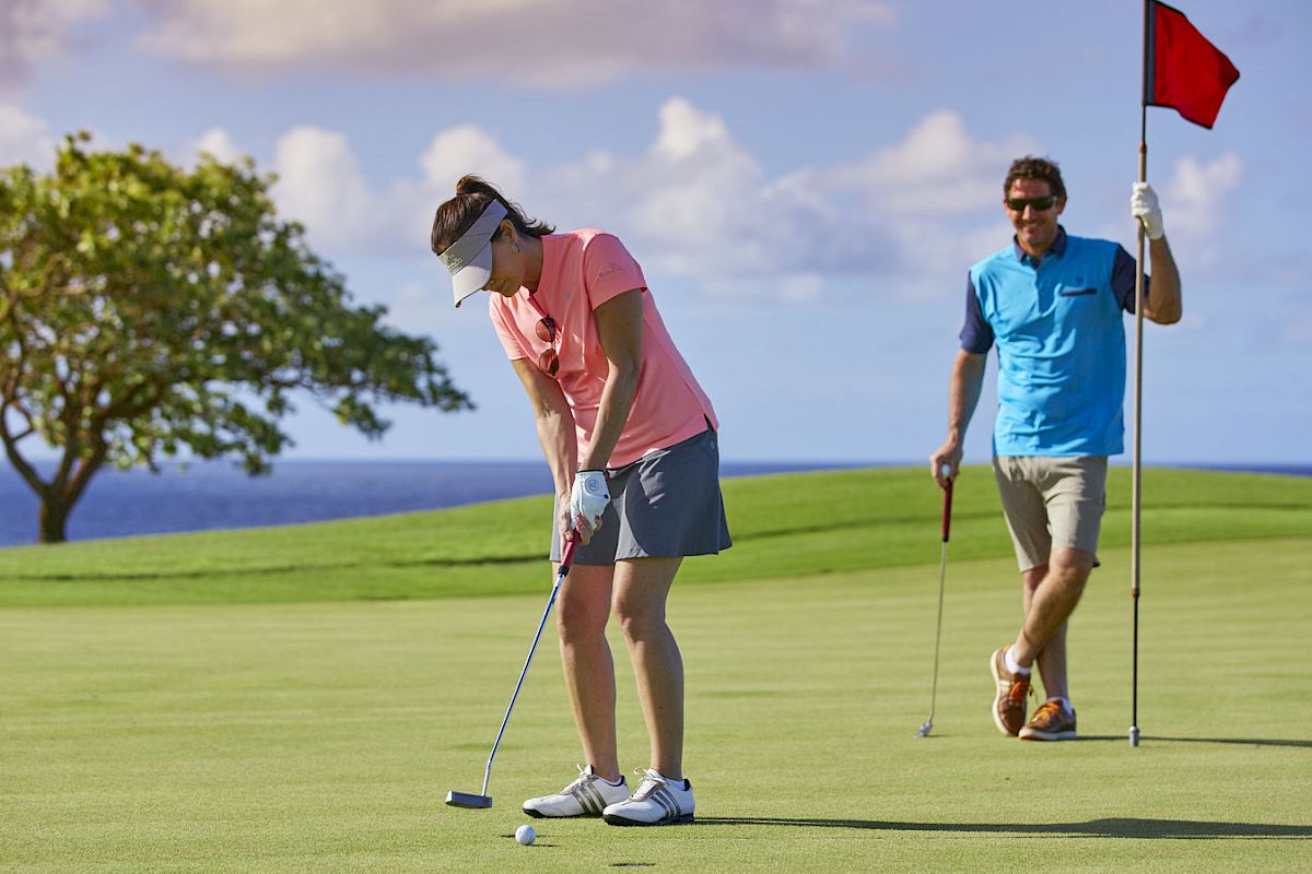 Two people are playing golf on a sunny day with a scenic ocean view. One is putting, and the other is holding the flag pin.