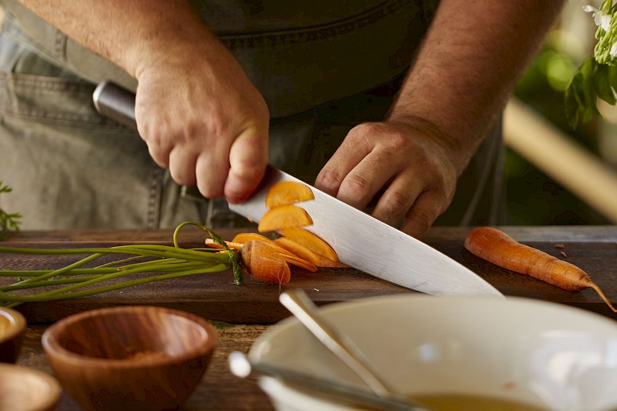 A person is slicing carrots on a wooden cutting board with various kitchen items around, including wooden bowls and a large white bowl.