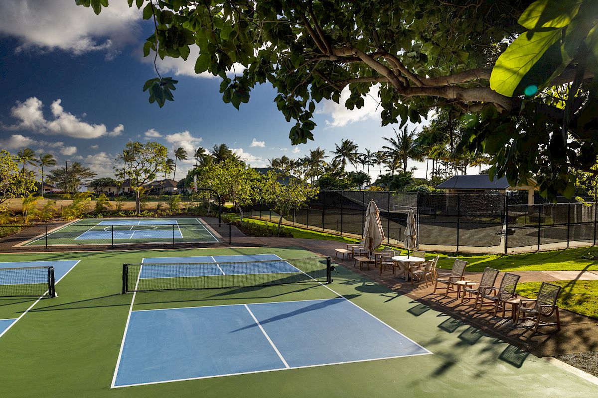 The image shows two outdoor tennis or pickleball courts under a clear sky, with chairs and umbrellas on the side, surrounded by greenery.