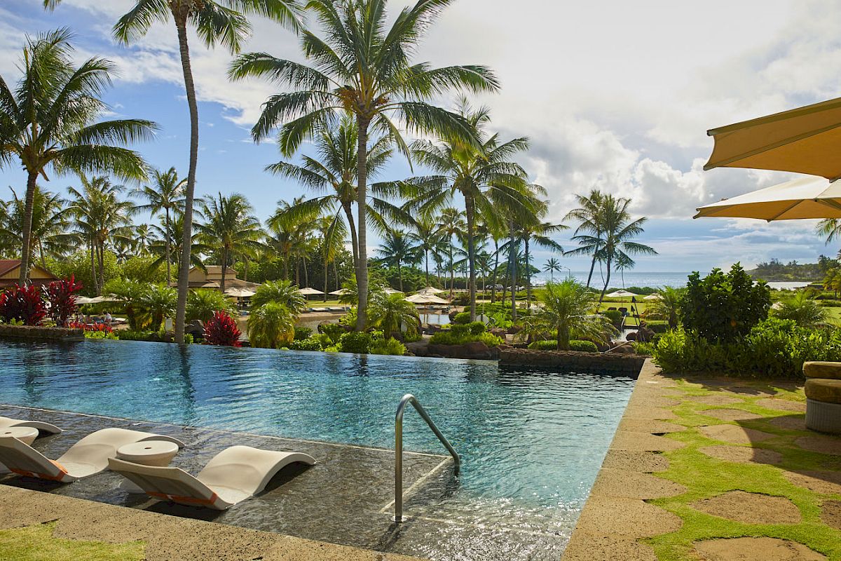 A serene poolside area with lounge chairs, surrounded by palm trees and greenery, with umbrellas providing shade.