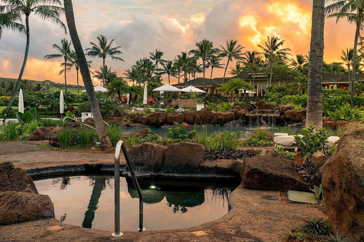 The image depicts a tropical resort scene with a hot tub, palm trees, lush vegetation, and a sunset sky in the background ending the sentence.