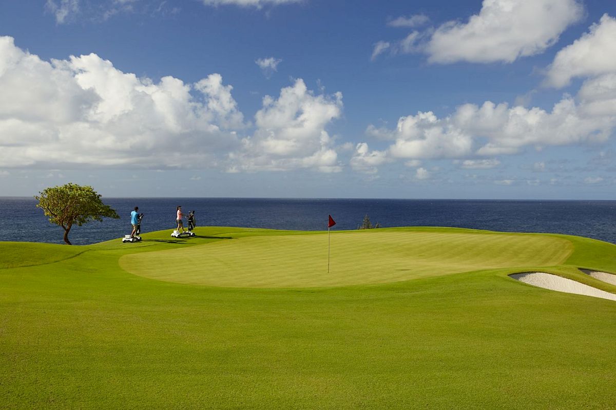 A scenic golf course with two people walking and a flag in the hole, set against a backdrop of the ocean and a partly cloudy sky.
