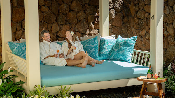 A couple relaxes on a cozy outdoor daybed, enjoying drinks and each other's company amidst lush greenery and stone walls.