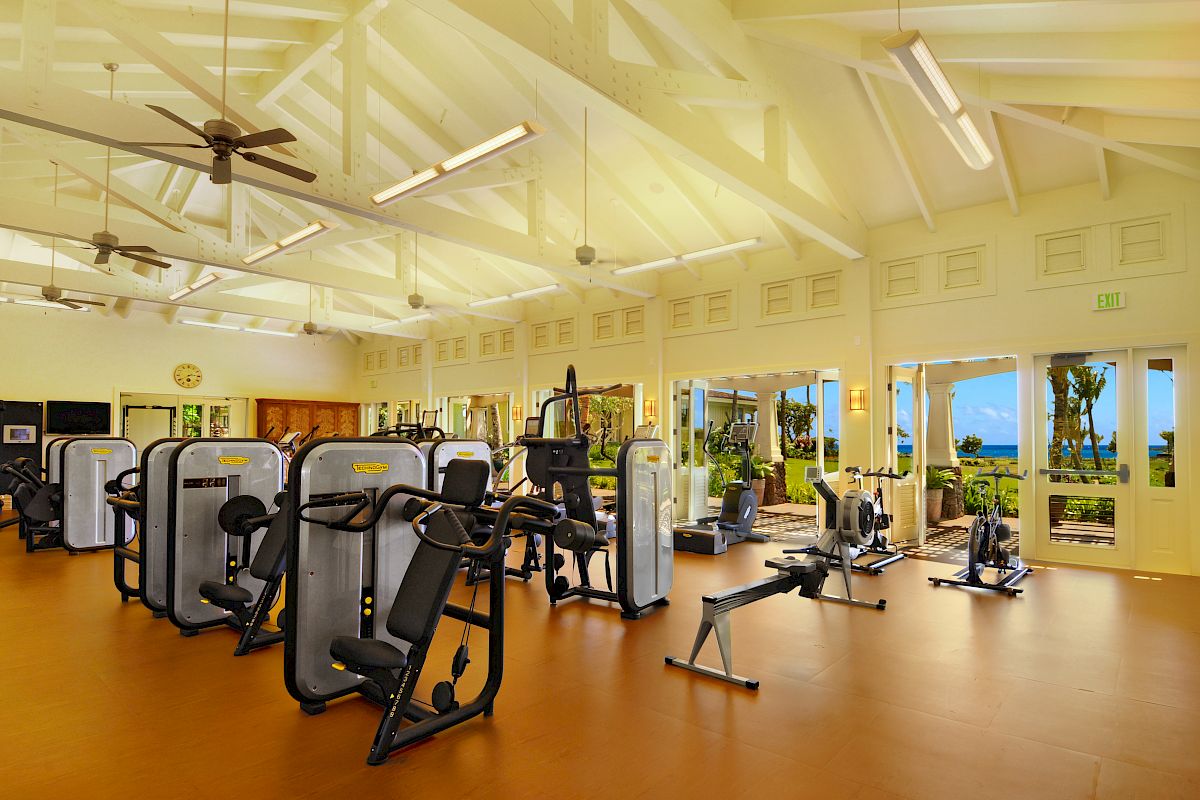 A spacious gym with various exercise machines, large windows for natural light, wooden floors, and a view of a tropical landscape outside.