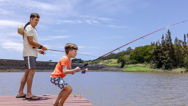 A man and a boy are fishing on a dock by a river, enjoying the outdoors on a sunny day. Excitement fills the moment.