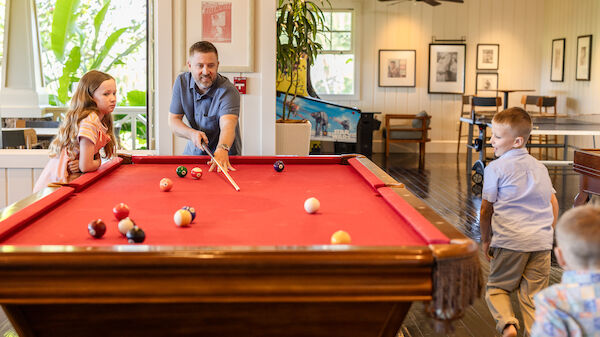 A man plays pool with two kids while a girl watches, in a bright room filled with plants and arcade games, creating a fun atmosphere.