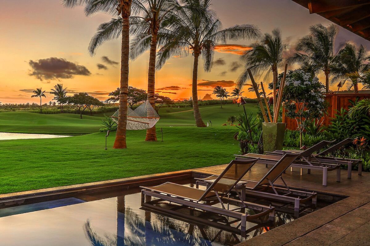 The image shows a peaceful outdoor setting with a pool, lounge chairs, palm trees, and a stunning sunset over green lawns.