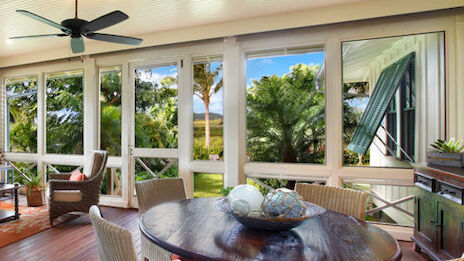 A bright, airy room with large windows, a ceiling fan, wicker chairs around a round table, and views of lush greenery outside.