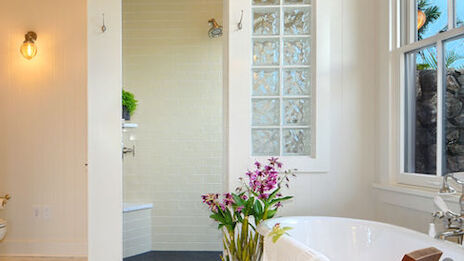 A modern bathroom with a freestanding bathtub, wooden chair, potted plants, and a shower area with glass bricks and light-colored tiles.
