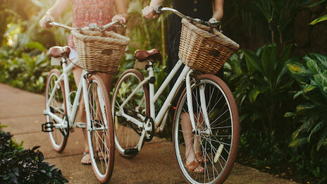 Two people are walking with their bicycles along a pathway, each bike equipped with a wicker basket, surrounded by lush greenery and sunlight.