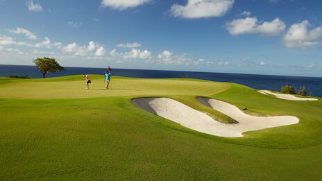 A picturesque seaside golf course with two people standing on the green, near a sand trap, under a clear blue sky with scattered clouds.