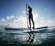 A person is paddleboarding on a calm sea under a clear sky, with the sun casting a silhouette on the water's surface.