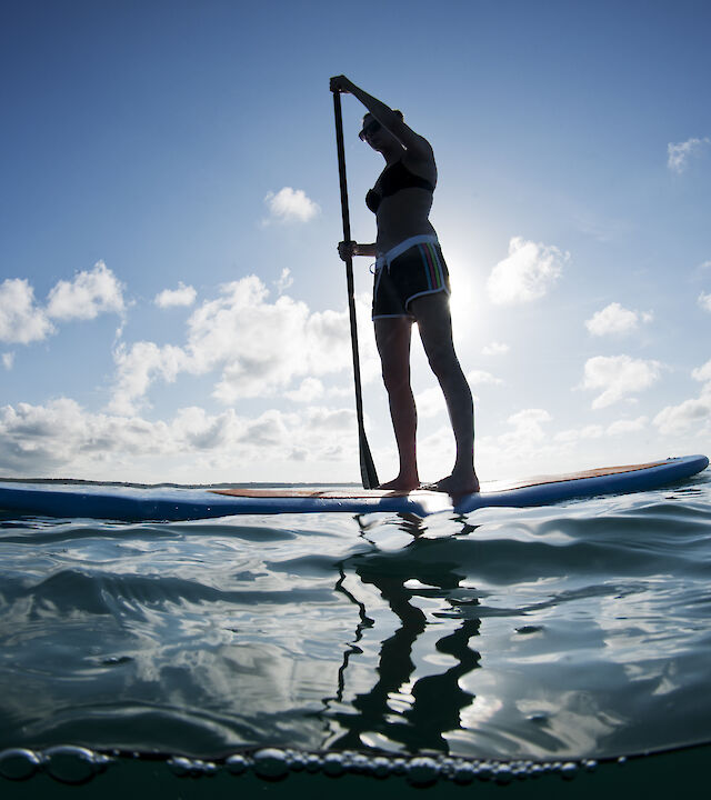 A person is paddleboarding on a calm sea under a clear sky, with the sun casting a silhouette on the water's surface.