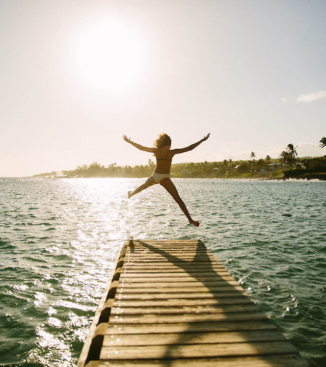 A person joyfully leaps from a dock into the water, illuminated by a bright sun with a serene landscape in the background.